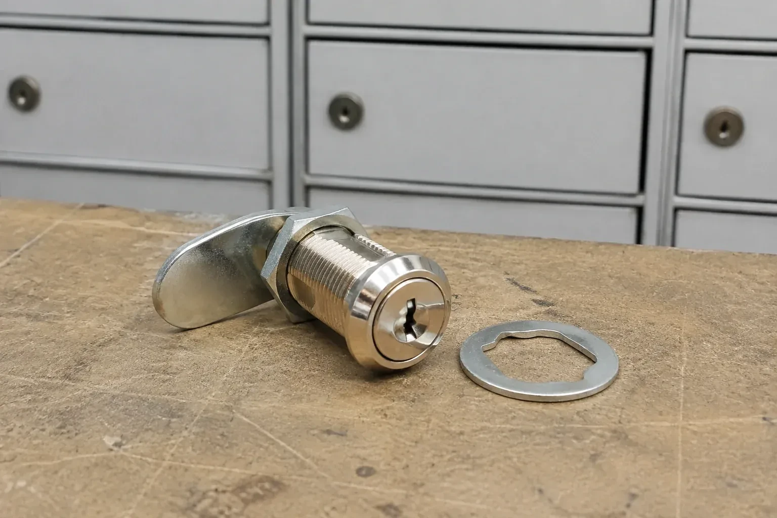 Detached threaded cam lock and mounting washer on a workbench in front of closed mail compartments. — Mailbox Lock Replacement, Aurora